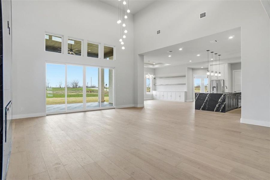 Expansive living room with soaring ceilings, wide-plank wood flooring, and sliding glass doors opening to the covered patio, seamlessly connecting the living area to the designer kitchen and dining space.
