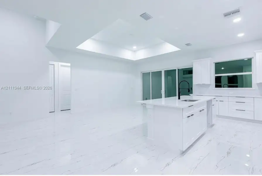 Kitchen featuring a central island with a quartz countertop, stone-finish tile flooring, recessed tray ceiling, white cabinetry with matte black hardware, and floor-to-ceiling glass sliding doors Kitchen featuring a central island with a quartz countertop, stone-finish tile flooring, recessed tray ceiling, white cabinetry with matte black hardware, and floor-to-ceiling glass sliding doors