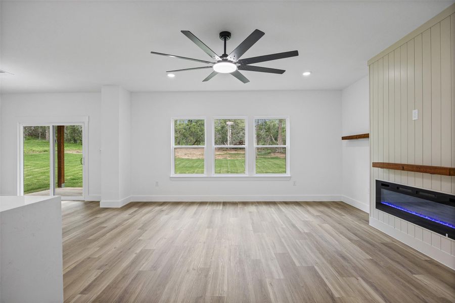 Spacious room featuring wood-finish flooring, a modern ceiling fan, recessed lighting, and a linear electric fireplace with wood mantel and shiplap-style accent wall Spacious room featuring wood-finish flooring, a modern ceiling fan, recessed lighting, and a linear electric fireplace with wood mantel and shiplap-style accent wall