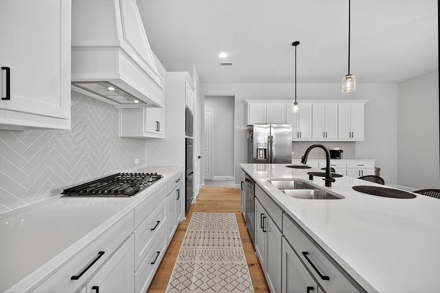 Gourmet kitchen featuring white shaker cabinetry, light-toned countertops, herringbone tile backsplash, stainless steel appliances, and a central island with a double basin sink and matte black fixtures