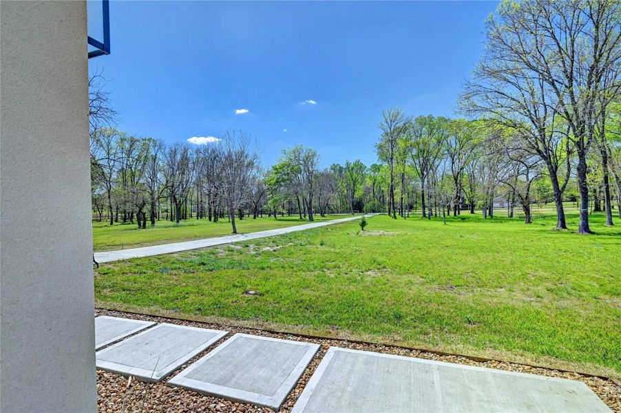 This photo shows a spacious, grassy landscape with scattered trees and a clear pathway, viewed from a patio area. The setting is peaceful and open, ideal for enjoying nature and outdoor activities.