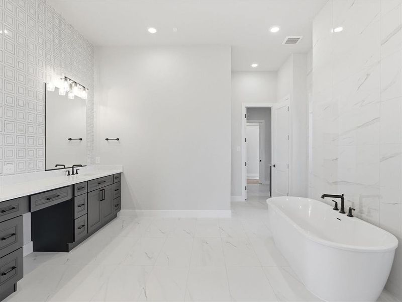 Spacious bathroom featuring a freestanding soaking tub, dual vanity with dark cabinetry and white countertops, large format floor tiling, and a tiled accent wall