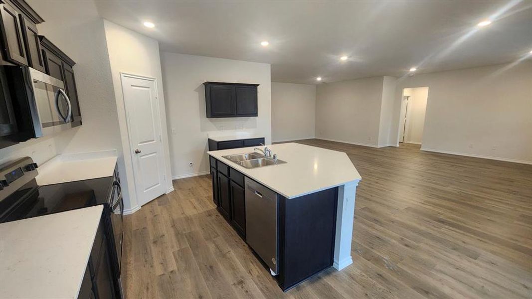 Open-concept kitchen featuring dark cabinetry, stainless steel appliances, and a central island with a dual sink and light-toned countertop