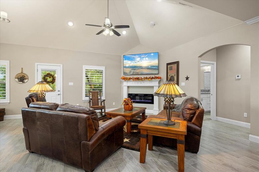 Living area featuring a glass covered fireplace, light wood-type flooring, vaulted ceiling, a ceiling fan, and arched walkways Living area featuring a glass covered fireplace, light wood-type flooring, vaulted ceiling, a ceiling fan, and arched walkways