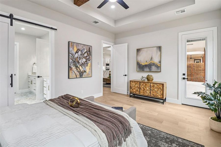 Bedroom with a barn door, ensuite bath, light wood-style floors, a tray ceiling, and ceiling fan