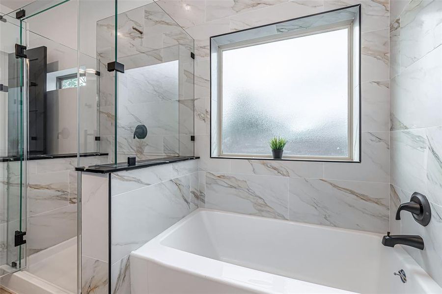 Modern primary bathroom featuring a combination of a tub and glass-enclosed shower. The room is adorned with marble-style tiles and includes a frosted window for privacy and natural light. Black fixtures add a contemporary touch to the space.