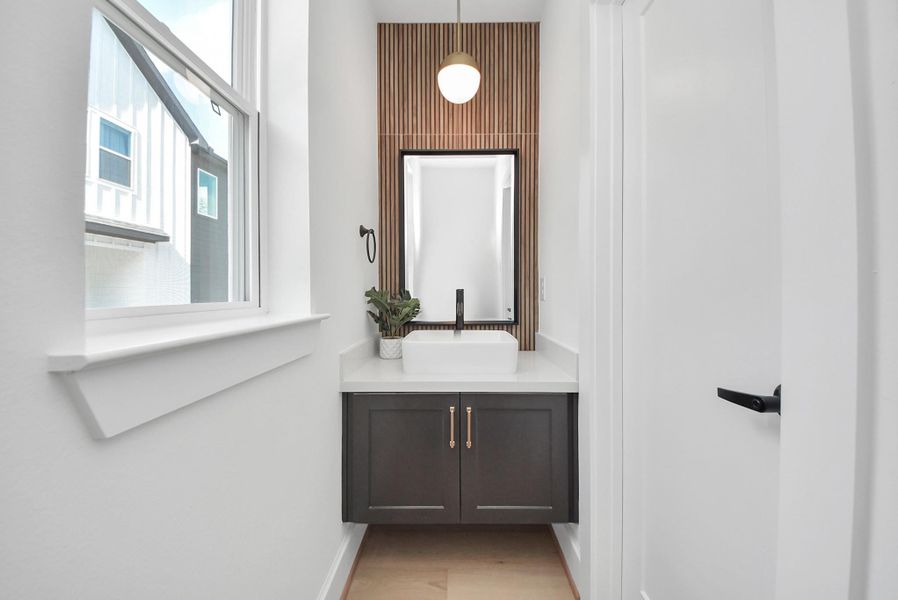 Step into this modern, compact bathroom featuring a sleek vanity with a white countertop and vessel sink. A stylish pendant light hangs above a wood-accent wall, while natural light from the window brightens the space, creating a clean and inviting design.