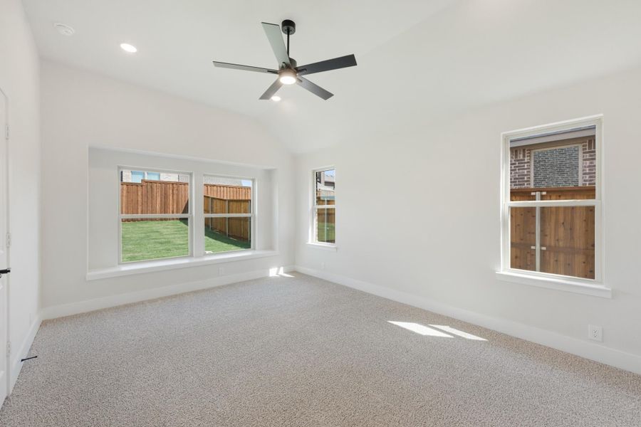 Empty bedroom with carpet flooring, ceiling fan, vaulted ceiling, and multiple windows overlooking a fenced backyard