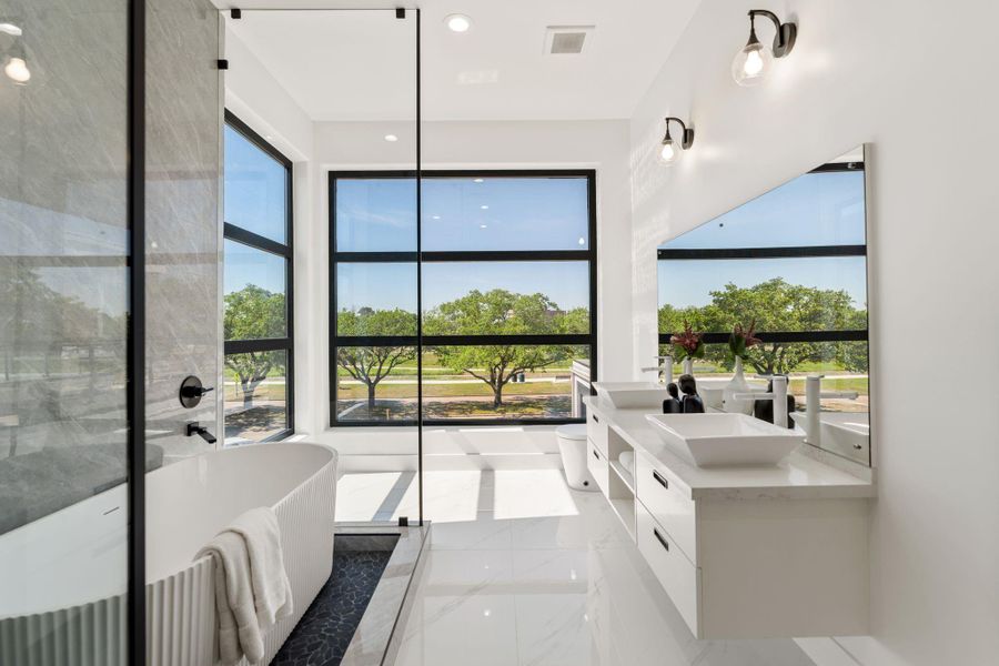 The freestanding soaking tub is positioned where it earns the most — at the floor-to-ceiling black-framed window wall, with a tree canopy view on one side and natural light pouring in from the other.