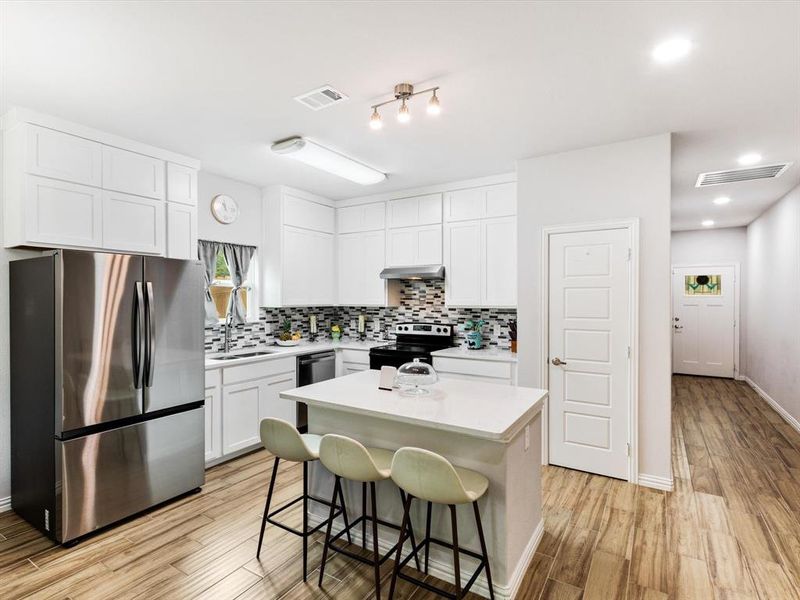 Bright kitchen featuring white cabinetry, a central island with countertop seating, stainless steel appliances, a mosaic tile backsplash, and wood-finish flooring