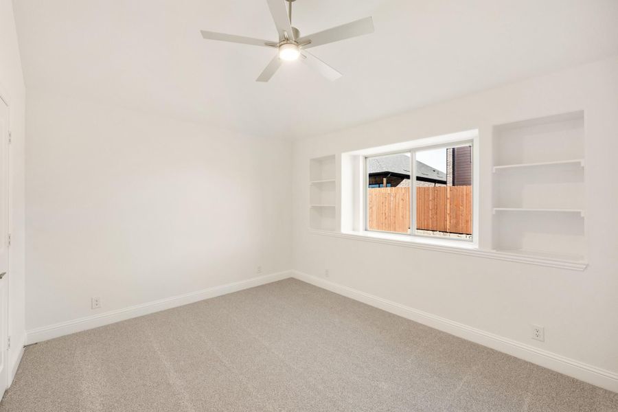 Empty bedroom with carpet, ceiling fan, built-in wall shelves, and a window overlooking a wood fence Empty bedroom with carpet, ceiling fan, built-in wall shelves, and a window overlooking a wood fence