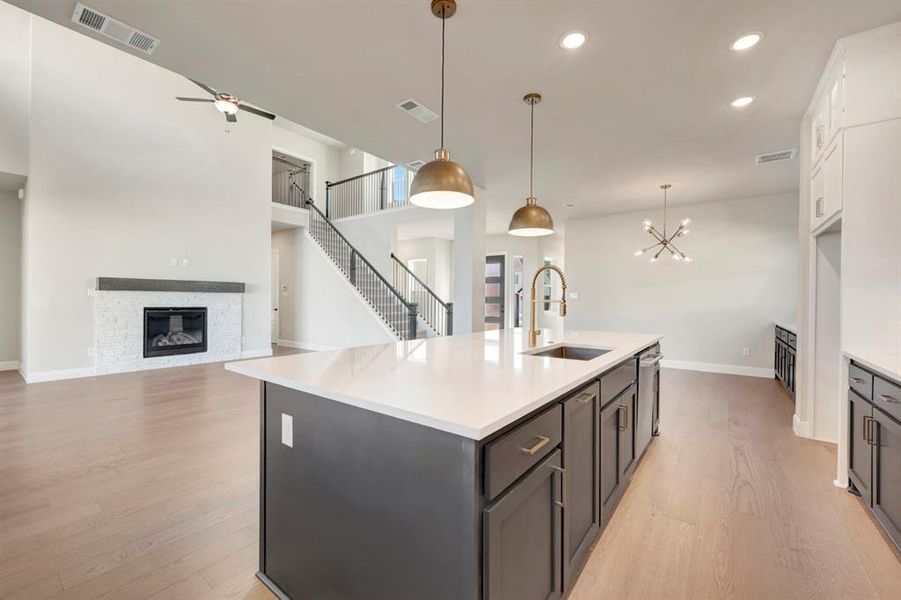 Kitchen featuring open floor plan, light wood-style flooring, a chandelier, a stone fireplace, and a kitchen island with sink Kitchen featuring open floor plan, light wood-style flooring, a chandelier, a stone fireplace, and a kitchen island with sink