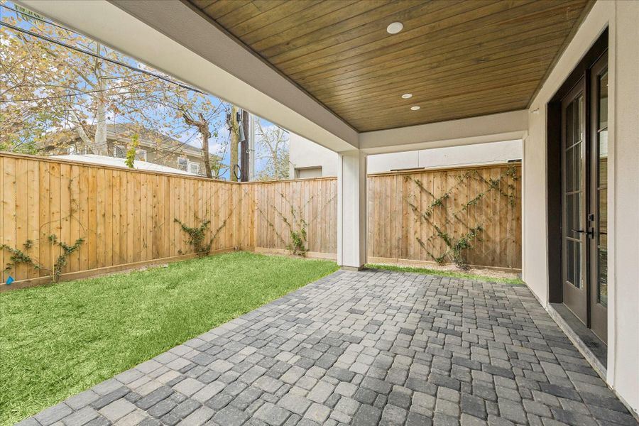 The first-floor Pavilion features a wood-plank ceiling with recessed lighting, cobblestone pavers, and lush lawn—a private outdoor retreat enclosed by cedar fencing with climbing vines. French doors connect seamlessly to the first-floor bedroom, whether used as a guest suite, office, or gym.