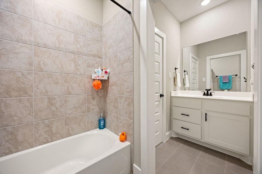 This bathroom features a sleek vanity with ample storage, complemented by modern hardware and a large framed mirror. Neutral tile flooring and a fully tiled tub/shower surround create a fresh and timeless look, offering both style and functionality.