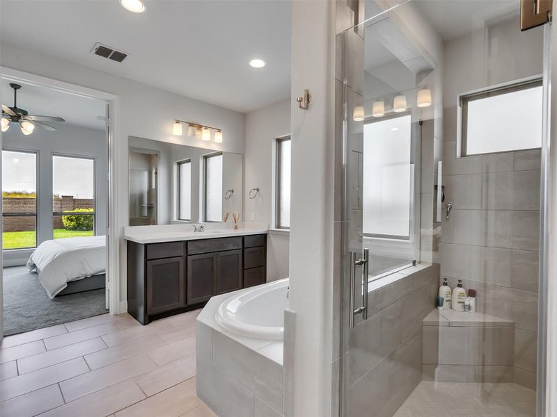 Experience luxury in this elegant bathroom featuring a spacious glass-enclosed shower, a relaxing soaking tub, and dual sinks with ample counter space. Adjacent, the bright bedroom offers serene views of the lush backyard.
