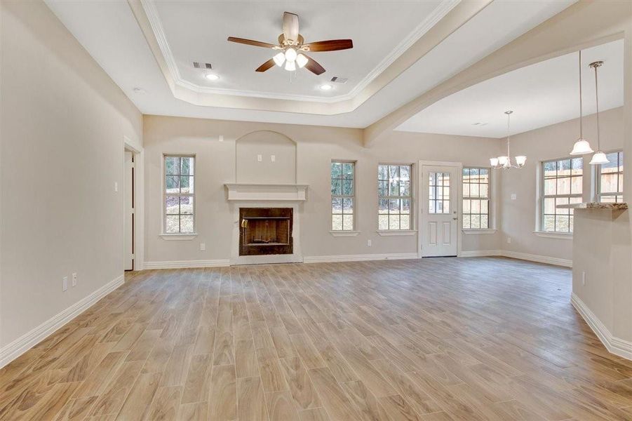 This spacious living area features a cozy fireplace, tray ceiling with fan, and abundant natural light from multiple windows. The open layout flows into a dining space, highlighted by elegant pendant lighting and wood-look flooring. This spacious living area features a cozy fireplace, tray ceiling with fan, and abundant natural light from multiple windows. The open layout flows into a dining space, highlighted by elegant pendant lighting and wood-look flooring.