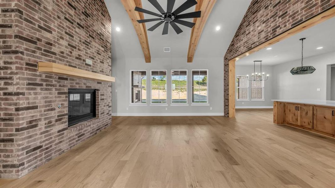 Vaulted great room featuring a brick fireplace with a wood mantel, exposed wood beams, wood-finish flooring, recessed lighting, and multiple windows Vaulted great room featuring a brick fireplace with a wood mantel, exposed wood beams, wood-finish flooring, recessed lighting, and multiple windows
