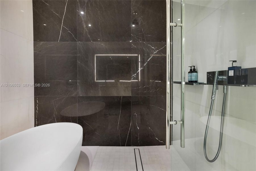 Freestanding soaking tub, floor-to-ceiling dark stone-finish wall tile, and glass-enclosed shower with polished chrome fixtures and an integrated shelf