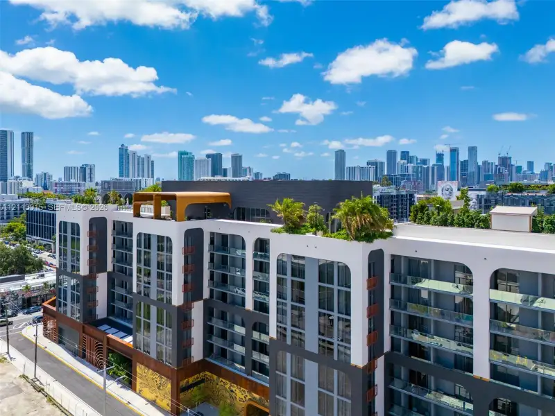 Modern multi-story building facade featuring arched architectural details, private glass-railed balconies, a rooftop terrace with mature landscaping, and multi-toned exterior paneling