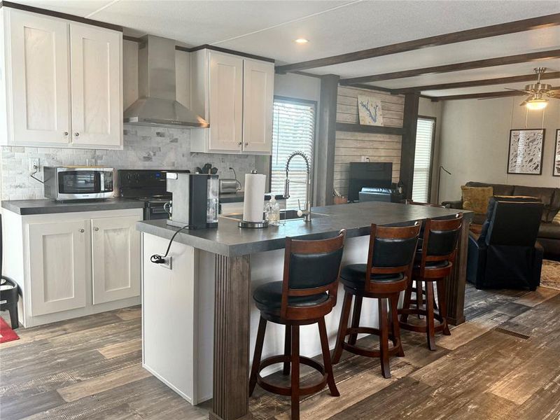 Kitchen with open floor plan, wall chimney range hood, dark countertops, dark wood finished floors, and beam ceiling