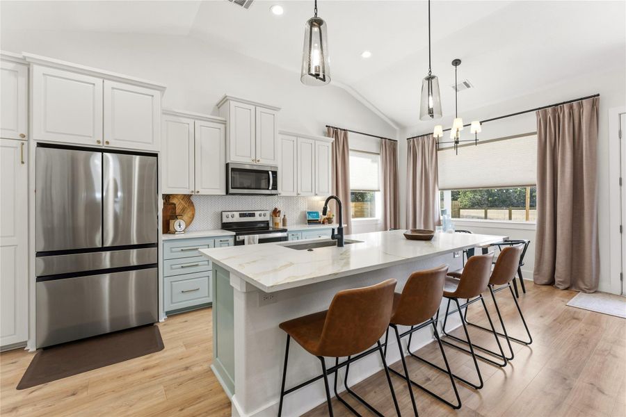 Two tone kitchen with stainless steel appliances, vaulted ceiling, light wood-style flooring, a kitchen island with sink, and suspended lighting
