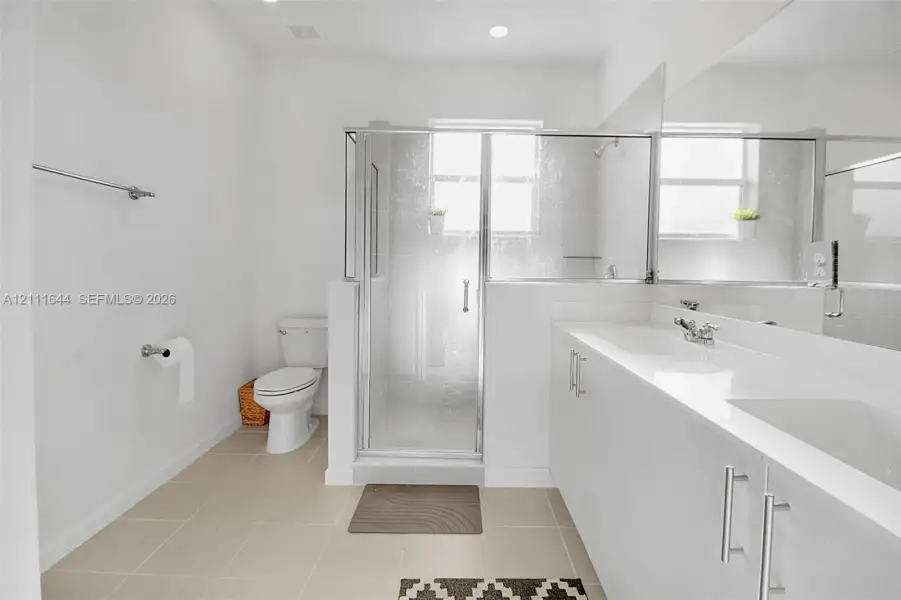 Dual-sink vanity with white cabinetry and solid surface countertop, glass-enclosed shower, polished chrome fixtures, and neutral floor tiling Dual-sink vanity with white cabinetry and solid surface countertop, glass-enclosed shower, polished chrome fixtures, and neutral floor tiling