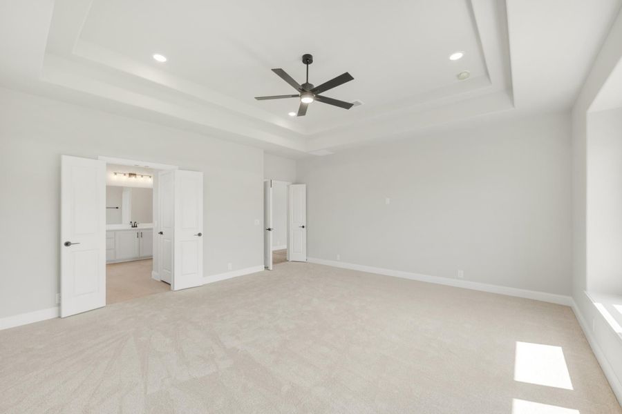 Spacious bedroom with tray ceiling, ceiling fan, carpet flooring, and doors leading to ensuite bathroom and closet. Spacious bedroom with tray ceiling, ceiling fan, carpet flooring, and doors leading to ensuite bathroom and closet.