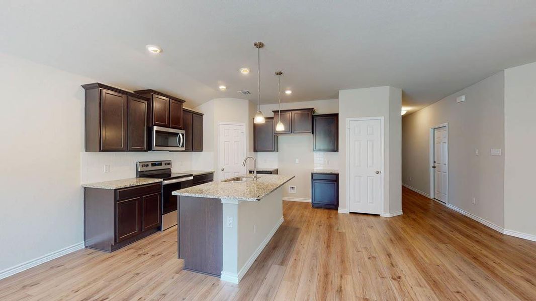 Kitchen featuring stainless steel appliances, dark wood finish cabinets, light stone countertops, hanging light fixtures, and a center island with sink