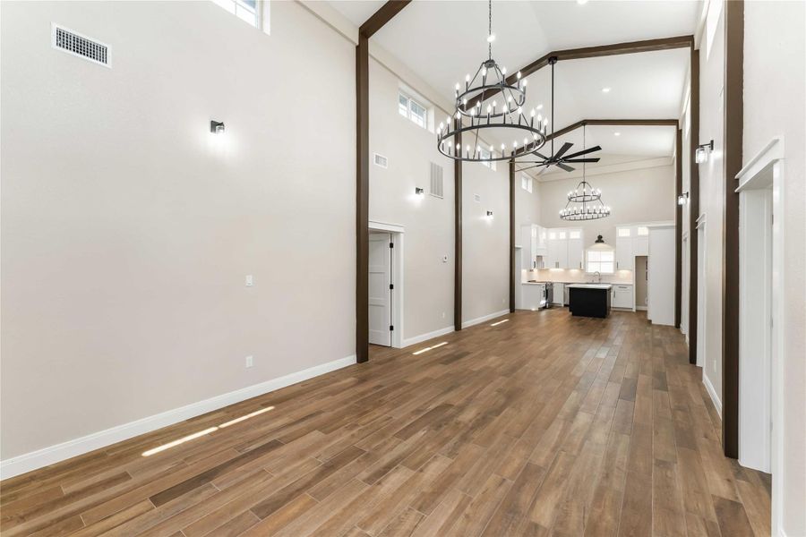 This spacious room features high ceilings with elegant chandeliers and exposed beams, complemented by warm tile wood-style flooring. It leads into a modern kitchen with white cabinetry and an island, offering an open, airy feel perfect for both entertaining and everyday living.