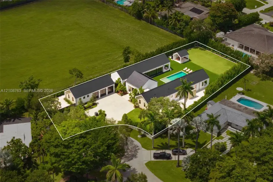 Estate featuring a multi-structure layout with metal roofing, a swimming pool, detached gazebo, expansive paved motor court, and manicured grounds enclosed by privacy hedges