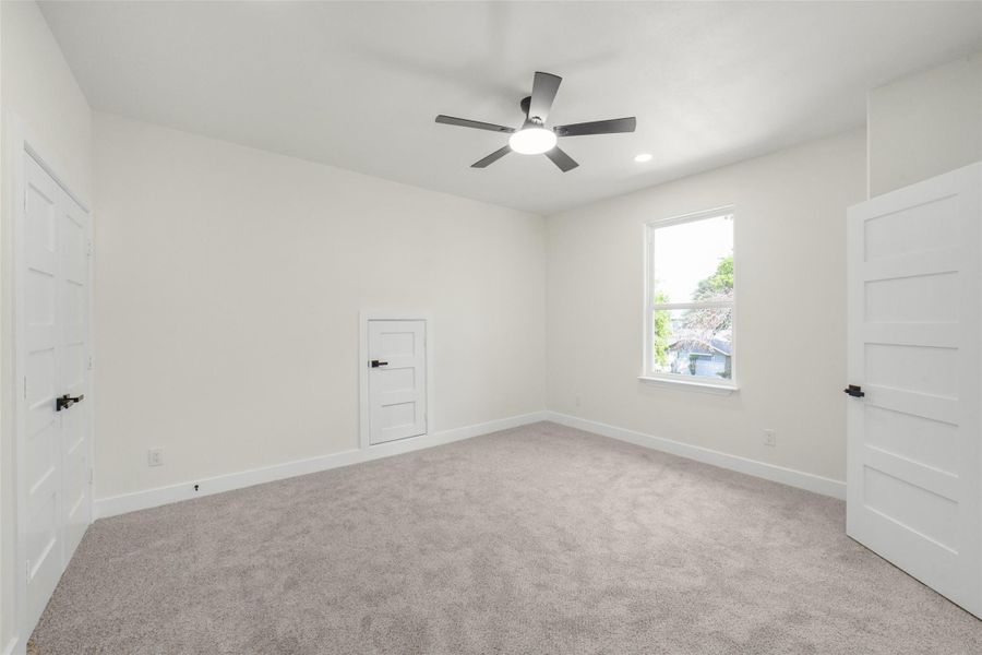 This bright, spacious room features neutral carpeting, a modern ceiling fan, and ample natural light from a large window. It includes clean white walls and multiple closets with sleek doors, offering plenty of storage space.