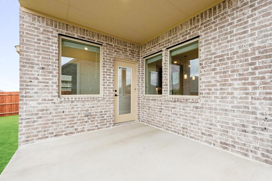 Covered back patio with concrete floor, light brick walls, back door, and two windows overlooking fenced yard