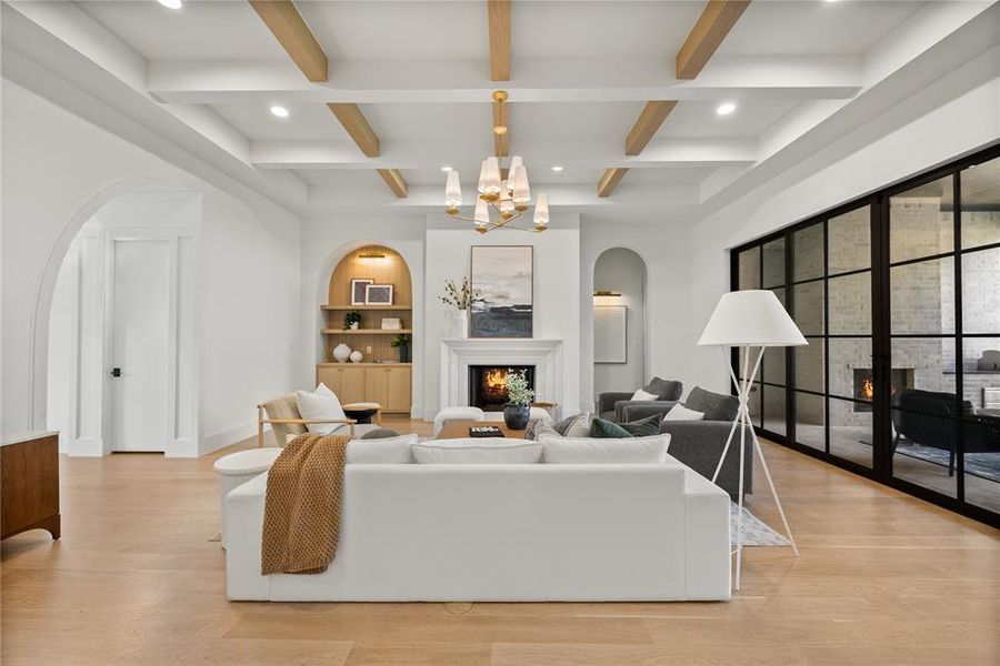 Spacious living area featuring a coffered ceiling with wood beams, light wood-finish flooring, a fireplace with a white mantel, arched doorways, and a wall of black-framed glass panels