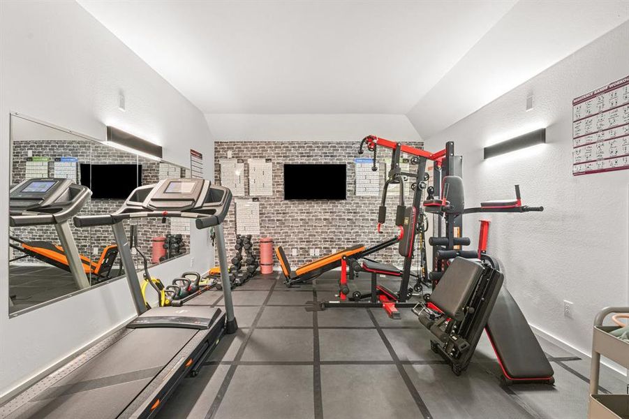 Workout area featuring vaulted ceiling, dark rubber floors, a textured wall, and brick wall