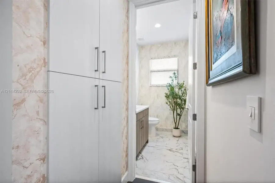 Floor-to-ceiling white cabinetry with brushed nickel hardware, natural stone-patterned tile flooring, wood-finish vanity, and recessed lighting