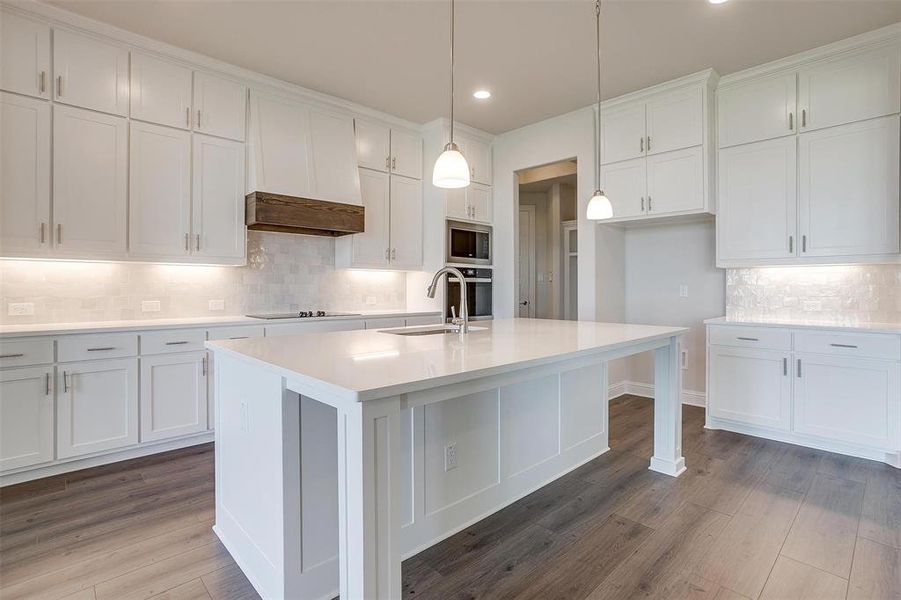 Kitchen with wood-type flooring, sink, stainless steel microwave, hanging light fixtures, and a center island with sink