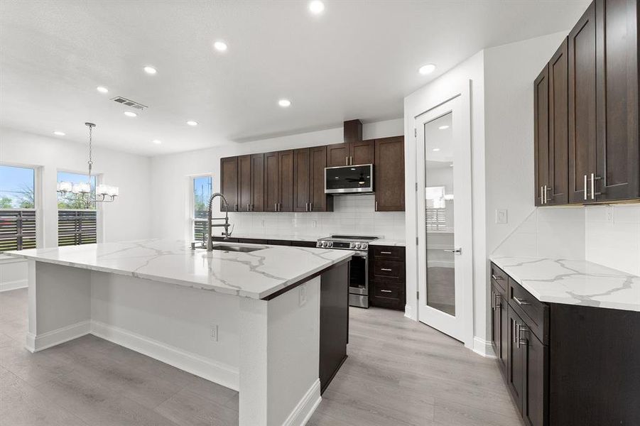 Kitchen featuring light stone countertops, decorative backsplash, stainless steel appliances, a center island with sink, and dark wood finish cabinetry
