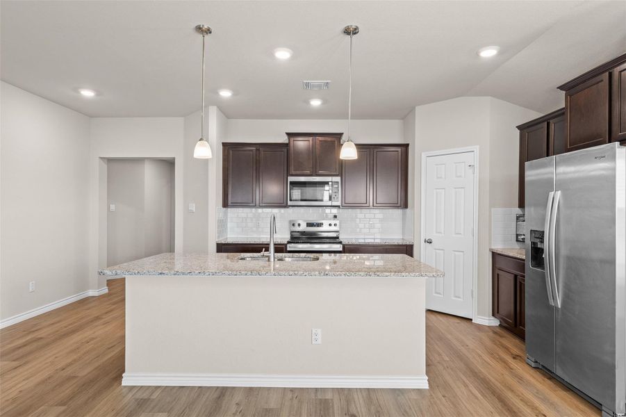 Kitchen with stainless steel appliances, dark wood finish cabinetry, light stone countertops, and a kitchen island with sink