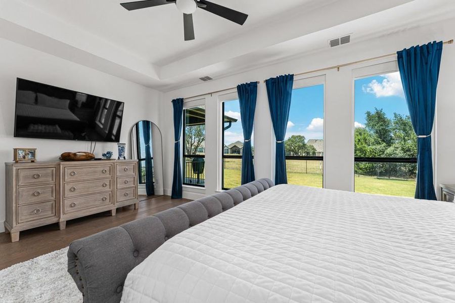 A spacious primary bedroom features a tray ceiling, wood flooring, and an abundance of natural light from large windows overlooking the backyard. A spacious primary bedroom features a tray ceiling, wood flooring, and an abundance of natural light from large windows overlooking the backyard.