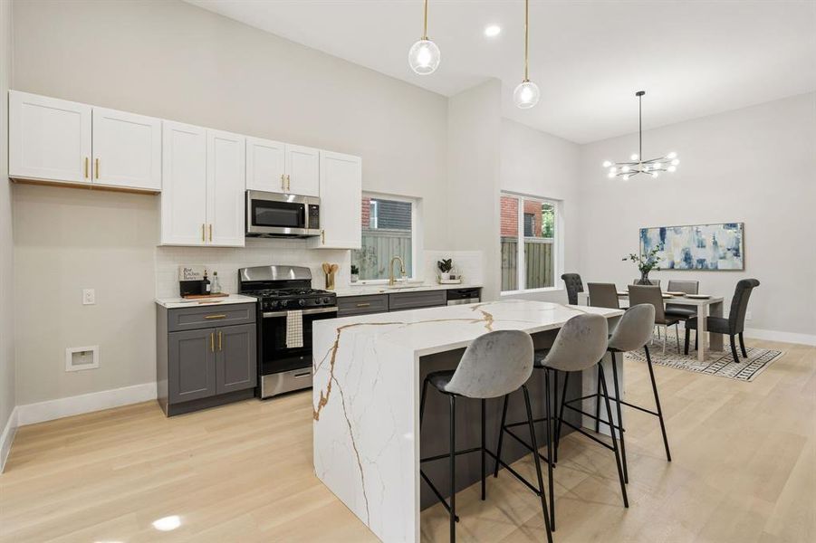 Open-concept kitchen featuring a waterfall island with a stone-finish countertop, two-tone cabinetry, stainless steel appliances, and light wood-finish flooring Open-concept kitchen featuring a waterfall island with a stone-finish countertop, two-tone cabinetry, stainless steel appliances, and light wood-finish flooring