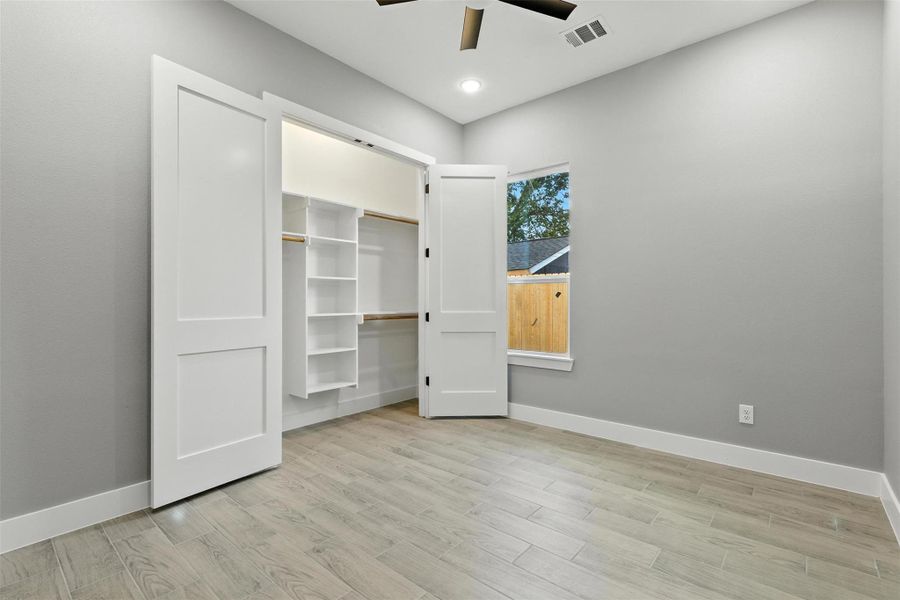 This room features light flooring and neutral gray walls, creating a bright and modern space. It includes a closet with sliding doors and built-in shelving. A window provides natural light and a view of a fenced yard.