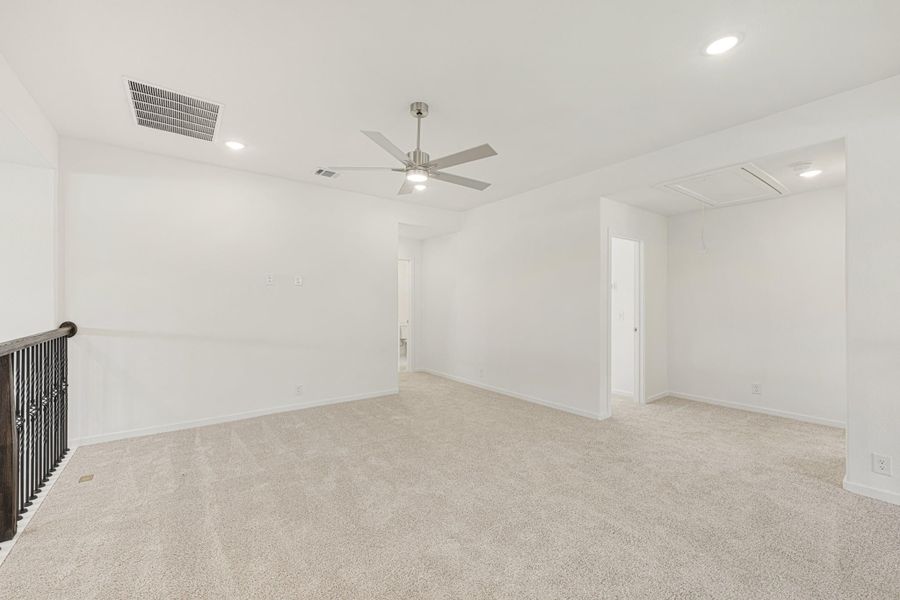 Open upstairs loft or bonus room with beige carpet, ceiling fan, white walls, and staircase railing visible