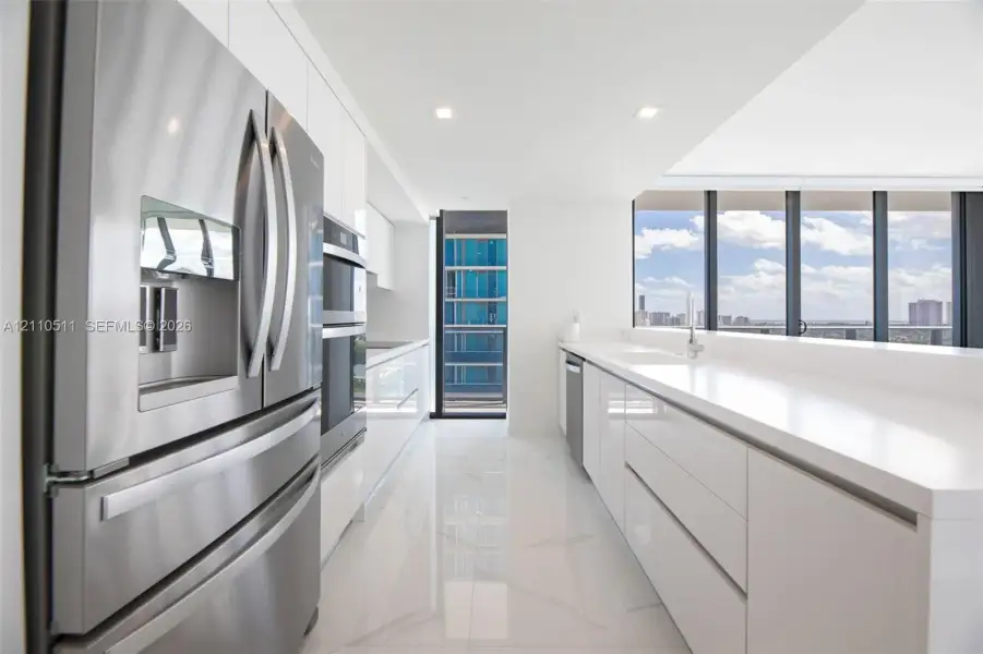 Modern kitchen featuring high-gloss white cabinetry, expansive stone countertops, stainless steel appliances, floor-to-ceiling windows, and large-format tile flooring