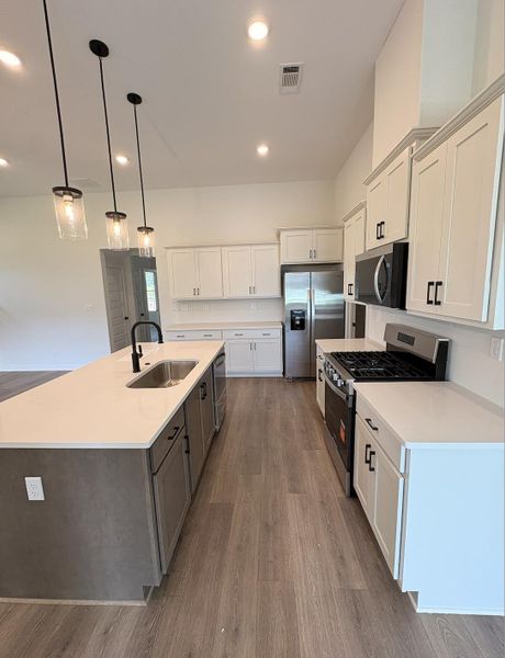Modern kitchen featuring an expansive center island with an undermount sink, white cabinetry with black hardware, wood-finish flooring, and stainless steel appliances Modern kitchen featuring an expansive center island with an undermount sink, white cabinetry with black hardware, wood-finish flooring, and stainless steel appliances