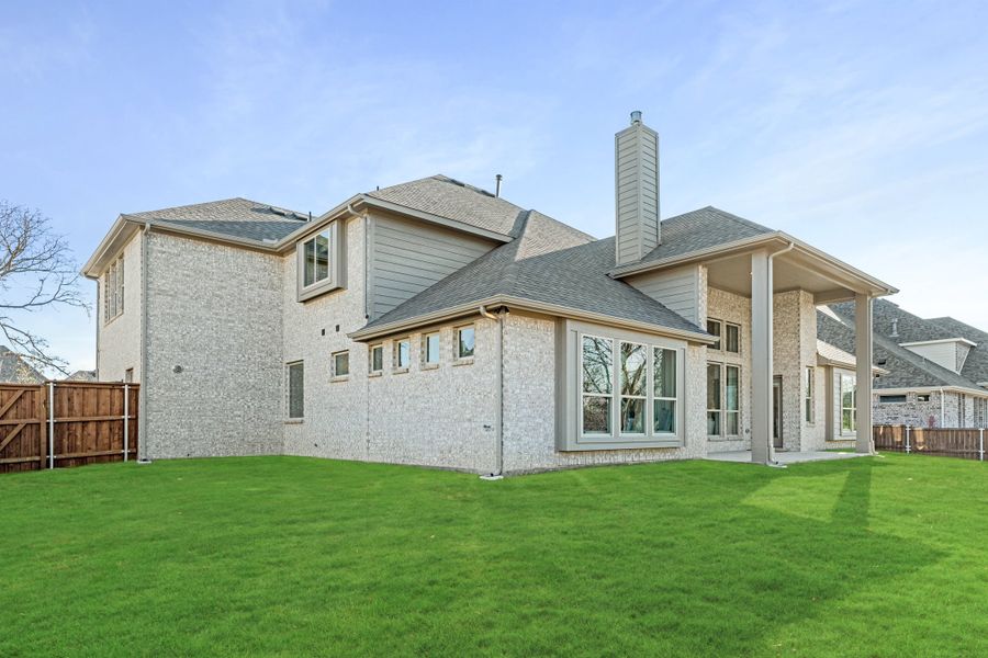 Two-story brick home rear exterior with chimney, covered patio, large windows, and fenced backyard with green lawn. Two-story brick home rear exterior with chimney, covered patio, large windows, and fenced backyard with green lawn.