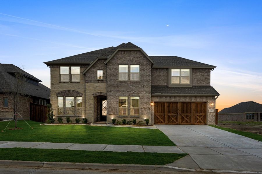 Two-story brick home with wood two-car garage doors, arched entry, and front lawn at dusk