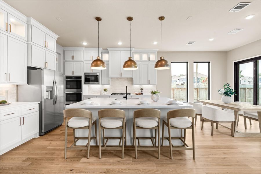 Elegant open-concept kitchen featuring sleek white cabinetry, a spacious island with modern pendant lighting, stainless steel appliances, and warm hardwood floors, seamlessly flowing into a bright living area with large windows and sliding glass doors. *Virtually Staged*