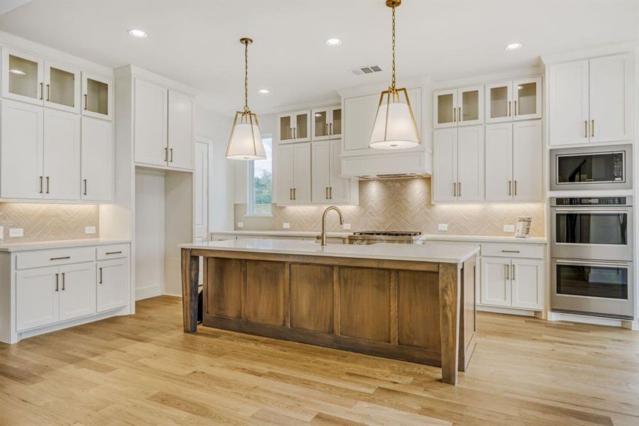 Kitchen with glass fronted cabinets, double oven, a center island with sink, light wood-style floors, and two tone cabinets