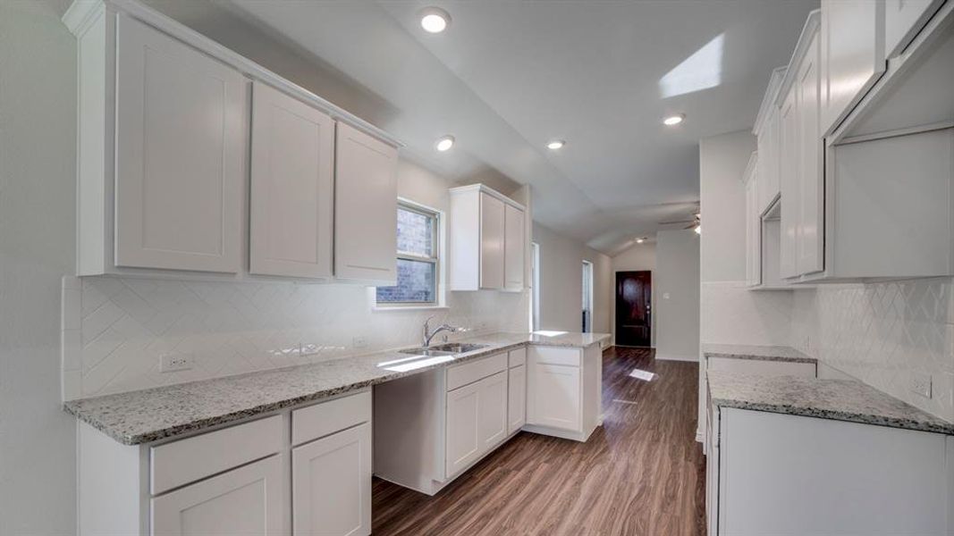 Kitchen featuring vaulted ceiling, tasteful backsplash, white cabinets, light stone counters, and dark wood-style flooring