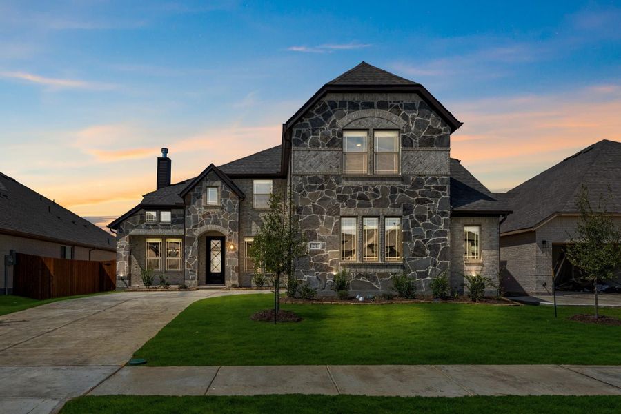 Two-story home with full stone exterior, arched entry, dark roof, and green front lawn at dusk